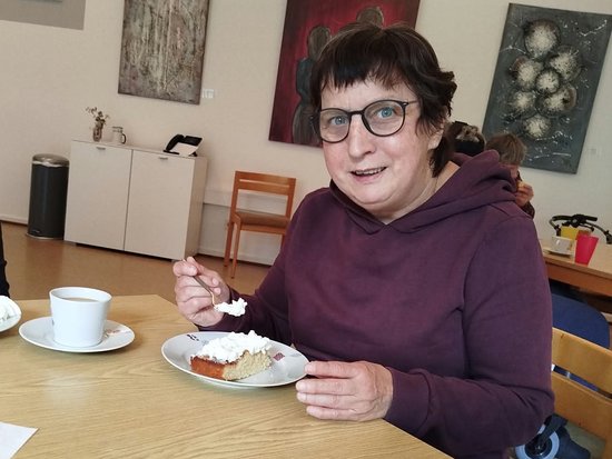 Eine Frau mit Brille und lila Kapuzenpullover sitzt an einem Holztisch und isst mit einer Gabel ein Stück Kuchen. Auf dem Tisch stehen eine weiße Tasse und ein blauer Becher. Im Hintergrund hängen Bilder an der Wand.