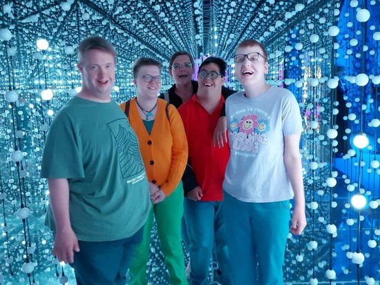 Fünf junge Männer stehen lächelnd nebeneinander in einem Spiegelraum. Der Raum ist mit vielen hängenden blauen und weißen Lichtern dekoriert, die einen unendlichen Effekt erzeugen. Die Spiegelwände reflektieren die Personen und die Lichter endlos.