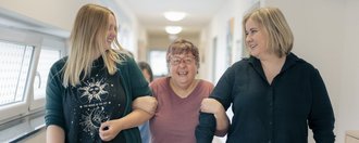 Haus Maria Helferin Zwei Mitarbeiterinnen gehen mit einer behinderten Frau fröhlich über den Flur