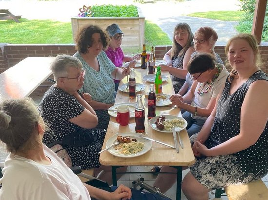 Eine Gruppe von Menschen sitzt an einem langen Holztisch unter einer Holzüberdachung und genießt gemeinsam ein Essen. Im Hintergrund ist ein grüner Garten zu sehen. Auf dem Tisch stehen Teller mit Speisen und Getränkeflaschen. Eine Person im Vordergrund sitzt im Rollstuhl.