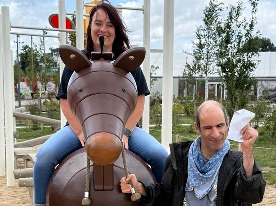 Eine Frau sitzt lächelnd auf einem braunen, tierförmigen Spielgerät. Ein Mann steht daneben und hält ein weißes Blatt Papier hoch. Beide wirken fröhlich auf einem sandigen Spielplatz.