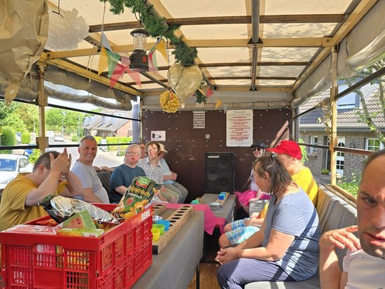 Eine Gruppe von Menschen sitzt in einem offenen, überdachten Fahrzeug und genießt eine Fahrt. Sie unterhalten sich, essen und trinken an Tischen. Im Vordergrund steht eine rote Kiste mit Snacks. Girlanden und bunte Flaggen hängen von der Decke des Fahrzeugs.