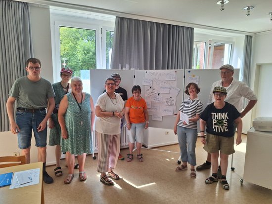 Eine Gruppe von neun Menschen steht in einem hellen Raum vor Whiteboards mit Notizen und Skizzen. Die Personen tragen lässige Sommerkleidung und posieren lächelnd für ein Gruppenfoto.